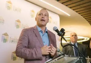 RC13 - St. Francis House Man in a pink blazer speaks at a podium with a microphone, in front of a backdrop with St. Francis House logos.