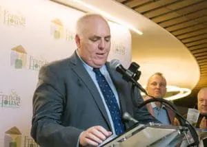 RC15 - St. Francis House Man in a gray suit and blue tie speaks at a podium with a microphone, St. Francis House backdrop.