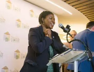 RC16 - St. Francis House African American woman in a navy blazer speaks at a podium with a microphone; backdrop shows St. Francis House logos at an event.