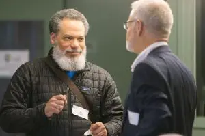 RC5 - St. Francis House Bearded man in a quilted jacket with a name badge talks to a colleague at a conference networking event.