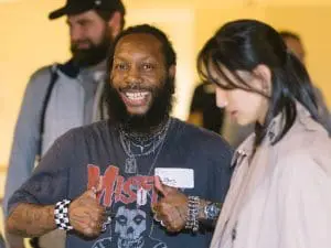 RC6 - St. Francis House Bearded man in a graphic tee with layered necklaces smiles and thumbs up at an indoor social event, standing beside a woman.