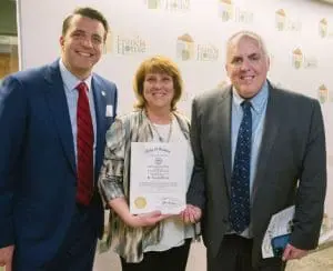 RC8 - St. Francis House Three adults in business attire pose for a photo; the woman in the center holds an award certificate, with a backdrop of St. Francis House logos.