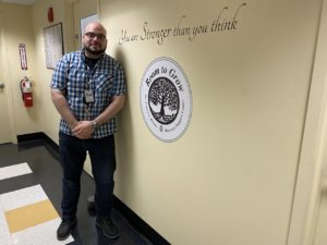 Jose Rodriguez stands in Recovery Support Center. He is leaning on a wall with that has a quote on it reading "You are Stonger thank you think." Below this quote is the Recovery Support Center's logo.