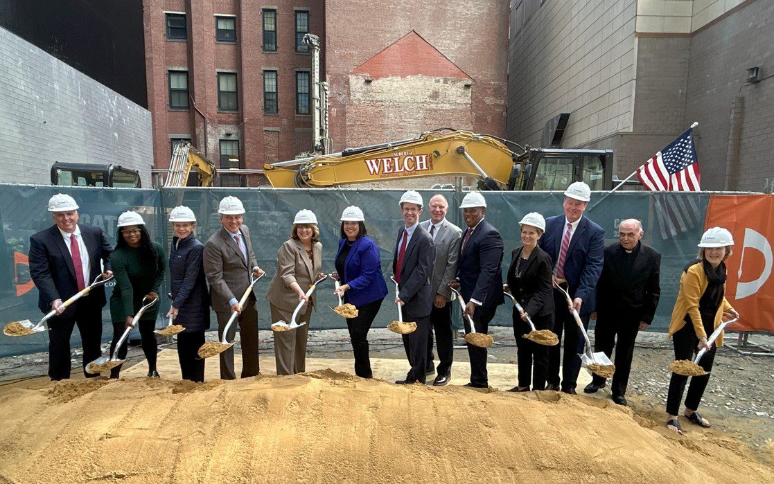 A line of dignitaries with hard hats and shovels on a dirt pile.