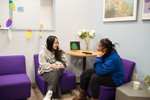 Two females sit in purple chairs in the Women's Center. They are smiling and chatting.