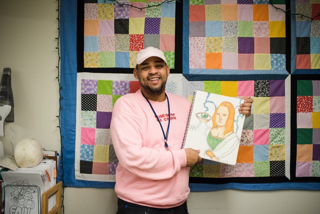 SFH Art Studio - St. Francis House A guest wearing a pink sweatshirt & hat stands in the art studio holding up one of his drawings.