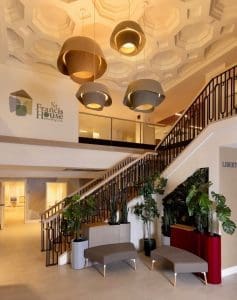 Bright lobby of St Francis House with large pendant lights, a sweeping staircase with a black railing, and potted plants.