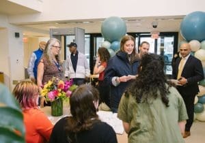 Reception desk where people are checking in for an event; a bouquet of flowers sits on the table and teal balloons frame the scene.