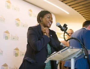 African American woman in a navy blazer speaks at a podium with a microphone; backdrop shows St. Francis House logos at an event.