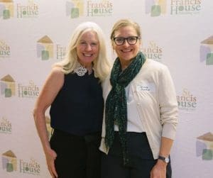 Two women smiling, standing side by side at a St. Francis House event; one wears black with a white lace collar, the other a white cardigan with a green scarf.