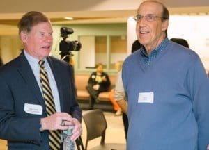 Two men in a conference setting chat; one in a suit with a striped tie, the other in a blue sweater, a camera in the background.