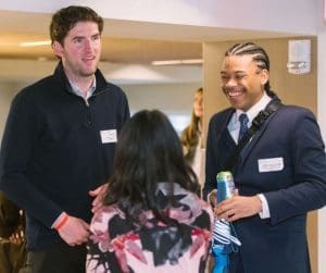 Two men and a woman converse in a hallway at a networking event; one man wears a navy suit with a name badge and drinks a can, smiling nearby.