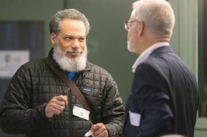 Bearded man in a quilted jacket with a name badge talks to a colleague at a conference networking event.
